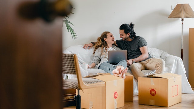 Smiling couple sitting on a couch looking at a laptop doing a free rental analysis surrounded by moving boxes